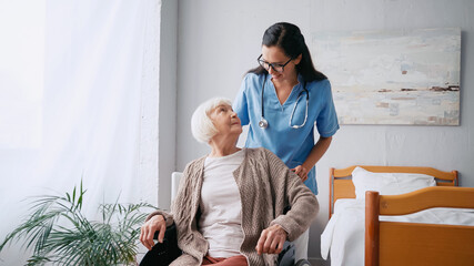 happy nurse in eyeglasses moving aged woman in wheelchair