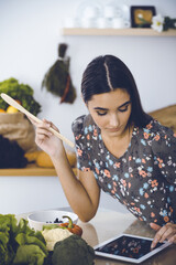 An attractive young dark-haired woman tastes a new recipe for a delicious salad mix while sitting at the table in the kitchen. Tablet pc is the best cookbook