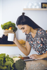 An attractive young dark-haired woman drinking morning coffee and looking for a new recipe for a delicious meal while sitting at the kitchen table. Tablet pc is the best cookbook