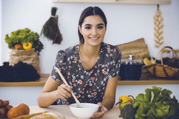 An attractive young dark-haired woman tastes a new recipe for a delicious salad mix while sitting and smiling in the kitchen. Cooking and householding concepts