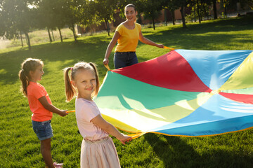 Group of children and teacher playing with rainbow playground parachute on green grass. Summer camp...