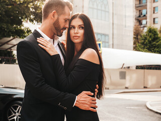 Beautiful fashion woman and her handsome elegant boyfriend in suit. Sexy brunette model in black evening dress. Fashionable couple posing in the street in Europe. Brutal man and his female outdoors