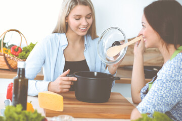 Two women friends looking into the dark pot with a ready meal and taste new recipes while sitting at the kitchen table. Healthy meal cooking concept