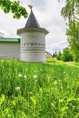 Low white stone fortress walls and a round fortress tower against the background of a green meadow with blooming dandelions on a sunny warm summer day