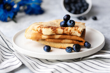 Tasty breakfast pancakes with blueberry meal on a white ceramic plate and flower bouquet in the background on a table striped linen cloth.