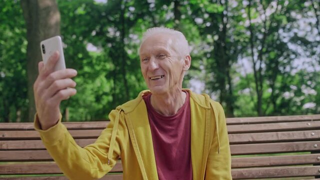 Mature man making video call to friends or relatives, sitting on bench in park