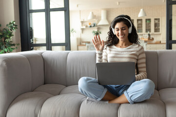 Young woman wearing headphones, communicating online on laptop, waving at webcam indoors, copy space