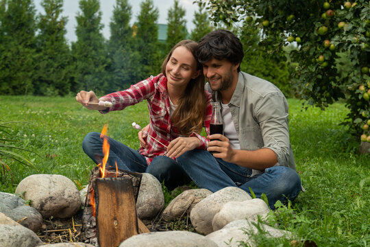 Pretty Couple Camping With Marshmallows Over A Campfire. Happy Young Couple Relaxing In The Backyard