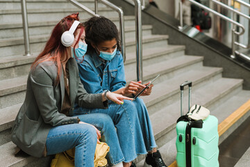 Multiracial two women in face masks sitting on stairs at train station