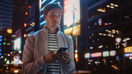 Portrait of Man Using Smartphone Walking Through Night City Street Full of Neon Light. Smiling Stylish Man Using Mobile Phone, Social Media, Online Shopping, Texting on Dating App.