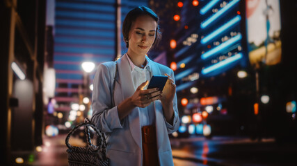 Beautiful Young Woman Using Smartphone Walking Through Night City Street Full of Neon Light. Smiling Thoughtfully Female Using Mobile Phone, Posting Social Media, Online Shopping, Texting.