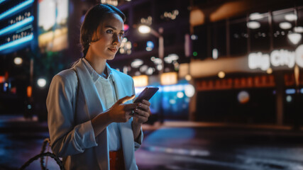 Beautiful Young Woman Using Smartphone Walking Through Night City Street Full of Neon Light. Portrait of Gorgeous Smiling Female Using Mobile Phone, Posting Social Media, Online Shopping, Texting