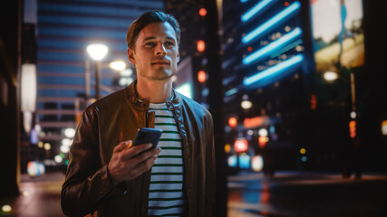 Man Using Smartphone Walking Through Night City Street Full of Neon Light. Smiling Stylish Man...