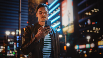 Man Using Smartphone Walking Through Night City Street Full of Neon Light. Smiling Stylish Man...