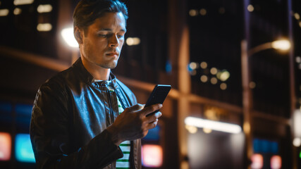 Portrait of Handsome Young Man Using Smartphone Standing in the Night City Street Full of Neon...