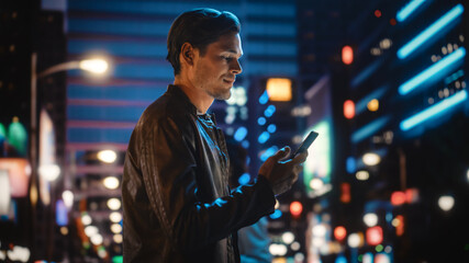 Portrait of Handsome Young Man Using Smartphone Standing in the Night City Street Full of Neon...