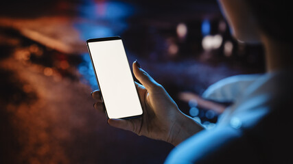 Beautiful Woman using Chroma Key Smartphone while Walking Through Night City Street Full of Neon Light. Female Using White Screen Mobile Phone. Over the Shoulder Close-up Shot - Powered by Adobe