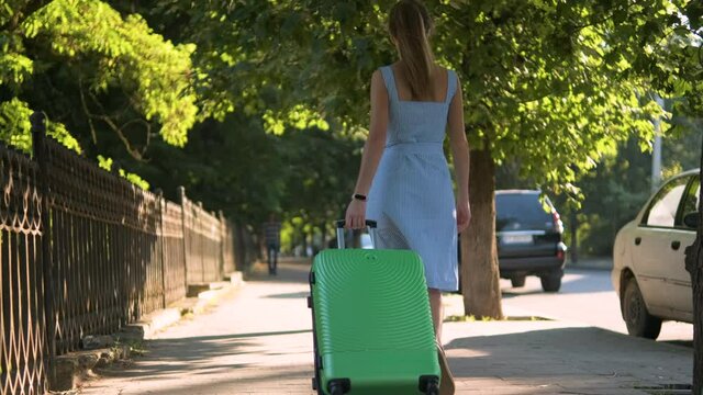 Young woman walking on city street sidewalk with green suitcase on summer day. Travelling and vacation concept.