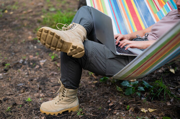 Caucasian woman working on laptop while sitting in a hammock in the forest. Girl uses a wireless computer on a hike.
