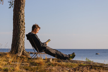 A man is reading a book sitting in camping chair outdoor.