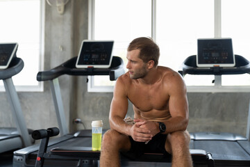 Muscular man sitting and drinking water after his workout in the fitness gym. Athlete drinking...