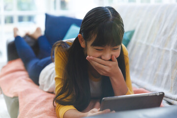 Smiling asian girl lying on couch in living room and using tablet - Powered by Adobe