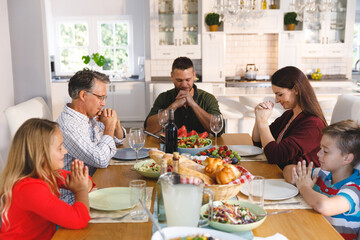 Caucasian grandfather and parents with son and daughter sitting at table and praying before dinner