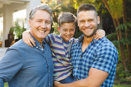 Portrait of smiling caucasian grandfather with adult son and grandson outside house in garden