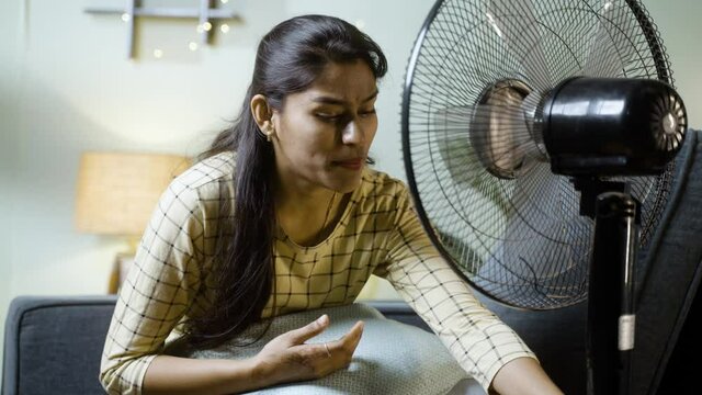 Indian Woman turning on fan due to summer heat wave while sitting on sofa - concept of heatstroke or feeling summer at home
