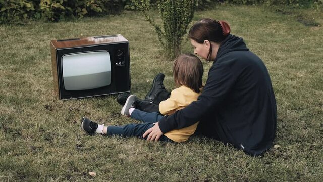 Woman, mother with small child are sitting on grass and watching an old retro TV. TV is strobing. Concept of post apocalypse, horror, surrealism and mysticism. Dependence on television