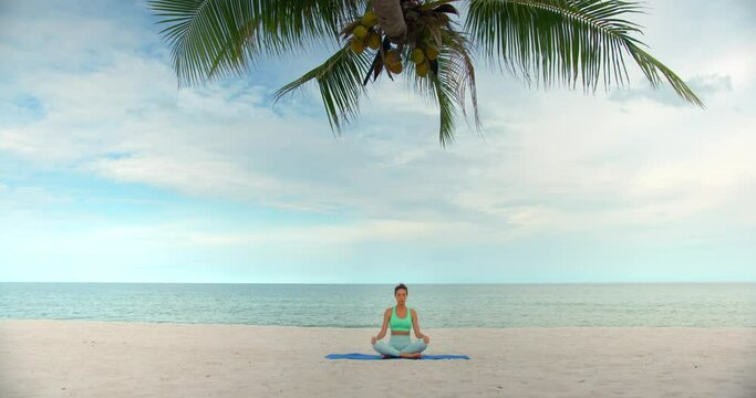 woman seats on yoga mat in lotus pose under the palm and meditating, she is located on the beach