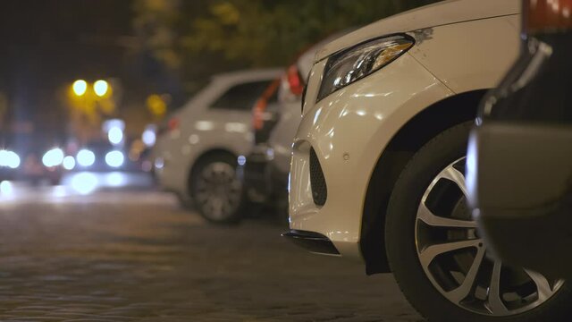 Close up of parked cars on roadside at night with blurred view of traffic lights of moving vehicles on city street.
