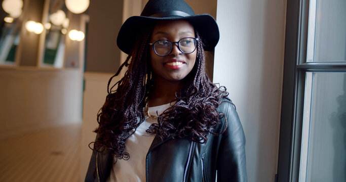 Portrait of stylish african woman in glasses and hat smiling standing indoors near window