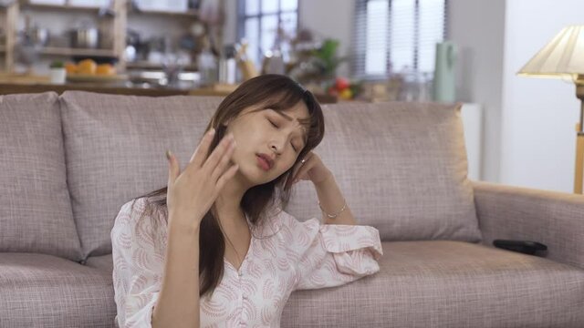 overheated asian woman is trying to cool down by fanning herself and pulling her clothes while feeling discomfort under high temperature near sofa at home.