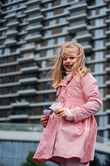 A cute, blond six-year-old girl in a pink coat stands in front of a tall building with a smile on her face and holds a flower in each hand.