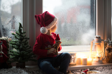 Beautiful toddler child, boy, waiting on the window on Christmas eve, looking for Santa Claus