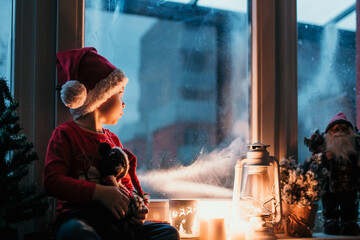 Beautiful toddler child, boy, waiting on the window on Christmas eve, looking for Santa Claus