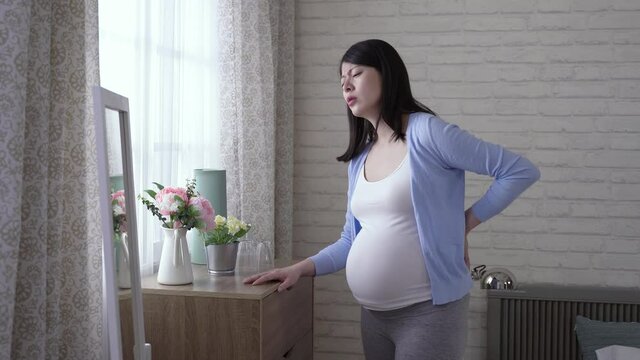asian lady in her last trimester is groaning in pain while trying to relieve the severe backache by self massage in the bedroom at daytime.