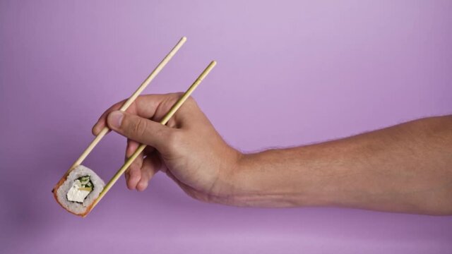 Close up on male caucasian hand holding wooden chop sticks with one roll sushi, isolated on puple background. Sushi restaurant, food delivery,  asian food, japanese cuisine delivery. 