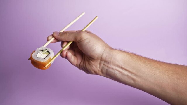 Close up on male caucasian hand holding wooden chop sticks with one roll sushi, isolated on puple background. Sushi restaurant, food delivery,  asian food, japanese cuisine delivery. 