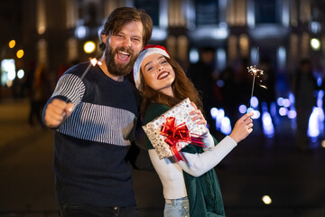 Young happy smiling couple walking at night city. A girl carries a gift box in her hand, a man holds a sparkler
