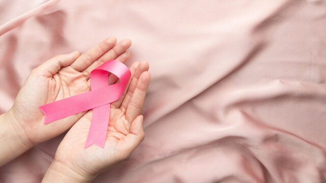 International symbol of Breast Cancer Awareness Month in October. Close up of female hand holding satin pink ribbon awareness on pink fabric background. Women's health care and medical concept.