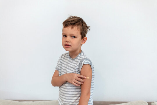 Adorable boy with adhesive bandage plaster on his arm after vaccination on grey background. Injection covid vaccine, healthcare for children and teenagers. Vaccinated boy showing arm after vaccination