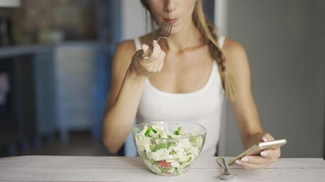 A happy woman is eating fresh vegetable salad and reading news or texting on smartphone. Slim woman is observing diet and counting calories. Concept of health or proper nutrition and weight loss.
