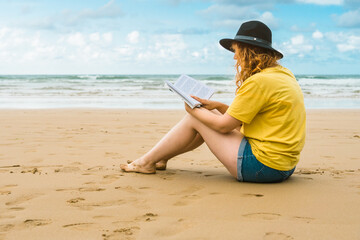 Closeup shot of a caucasian female in a yellow t-shirt reading a book at the beach