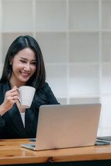 Portrait of young asian businesswoman hoding mug and working on laptop computer while sitting at her desk office.