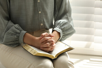 Religious woman praying over Bible near window indoors, closeup