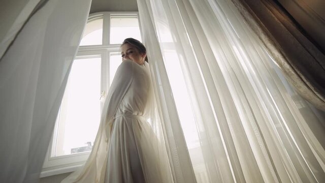 Bride in white boudoir dress dancing near window. Wedding morning preparations. Luxury bride with fashion hairstyle and makeup in a hotel or apartment. Beautiful, lovely woman in night gown and veil