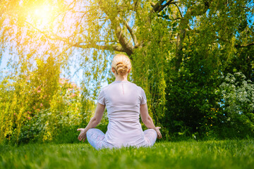 young woman doing yoga asana in park. girl stretching exercise in yoga position. happy and healthy woman sitting in lotus position and practicing yoga. meditation and sport on sunset outdoors 