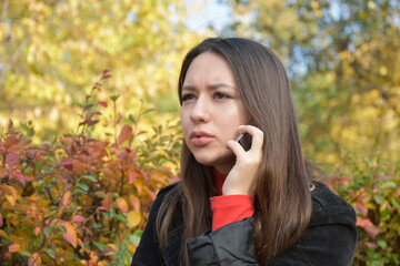 Serious young woman with long straight dark brown hair frowning and looking away while talking on phone in park in autumn on blurred background